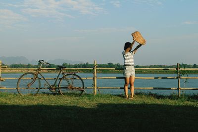 Rear view of woman standing by bicycle on field against sky