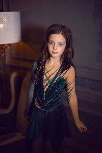 Girl in a green figure skating dress stands next to a chair and a lamp in the room