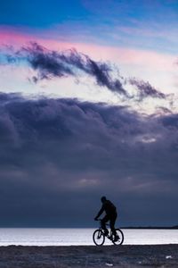 Man riding bicycle on sea against sky during sunset