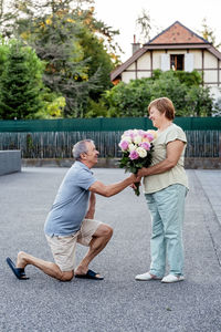 Happy smiling senior adult man giving bouquet of roses flowers to his mature wife standing on knee