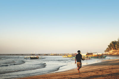 Rear view of man walking on beach by anchored boats