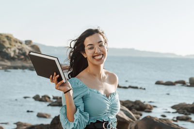 Portrait of smiling woman holding book against sea