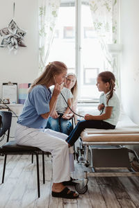 Pediatrician listening to girl's heartbeat through stethoscope while mother sitting with boy in background at clinic