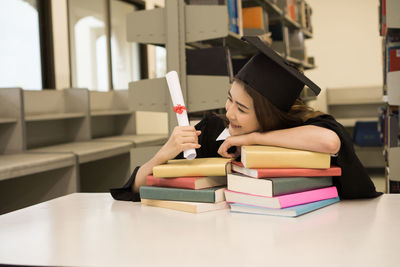 Young woman in graduation gown leaning on books at table in library