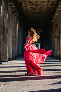 Full length side view of young woman in pink evening gown dancing at colonnade