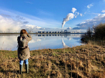 Rear view of woman watching industrial area with smoke stack at other side of mirror-like lake