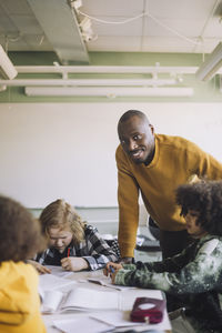 Portrait of smiling male teacher leaning at desk while students studying in classroom