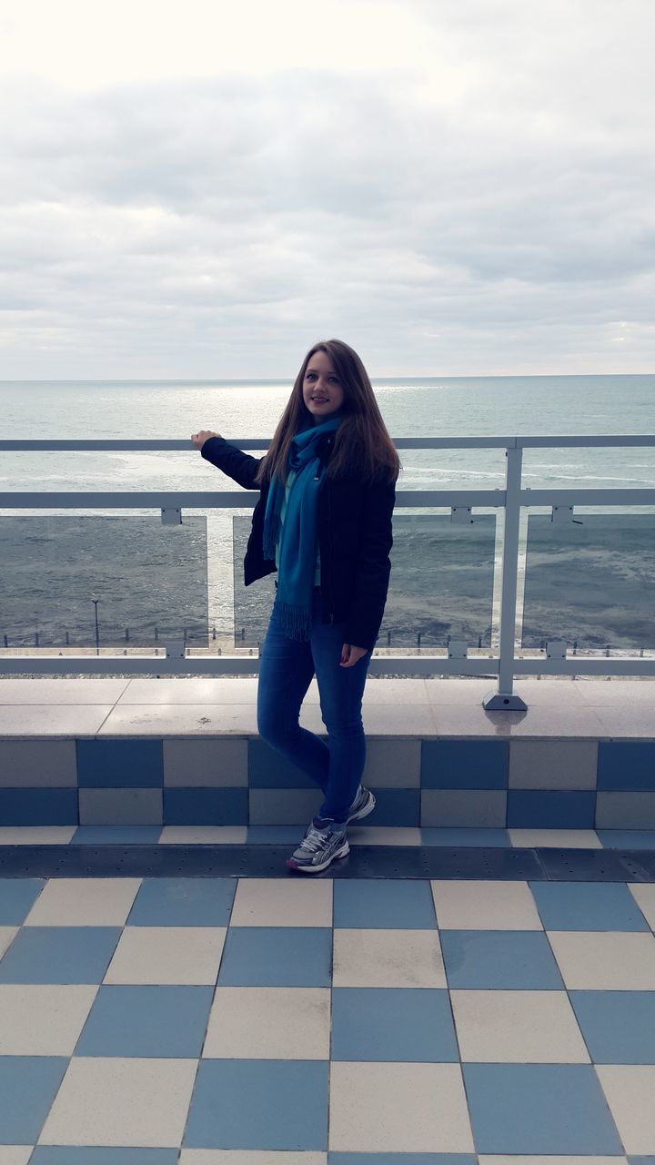 full length, sea, lifestyles, young adult, casual clothing, water, standing, person, leisure activity, sky, young women, horizon over water, portrait, beach, looking at camera, railing, front view