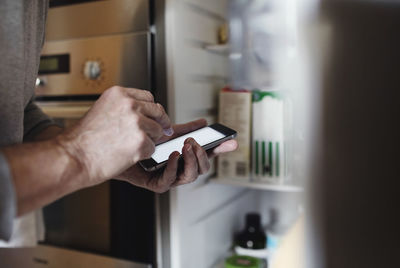 Cropped image of senior man using mobile phone while standing by open refrigerator