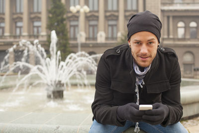 Young man using mobile phone while sitting against water fountain in city