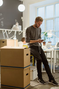 Full length of businessman using smart phone by cardboard boxes at office