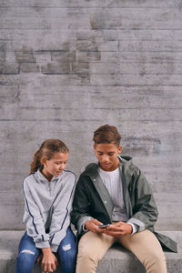 Siblings using mobile phone while sitting against wall