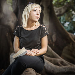 Young woman with book sitting on tree root at forest