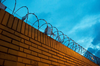 Low angle view of razor wire fence on brick wall against cloudy sky