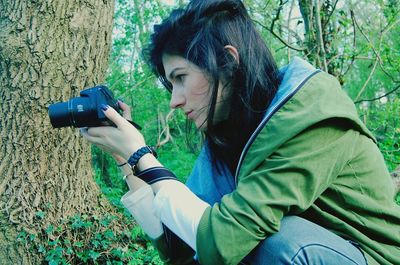 Side view of woman photographing through camera against trees at forest