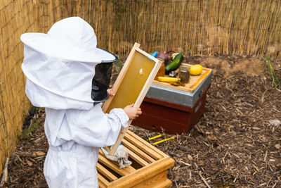 Side view of unrecognizable kid in protective costume standing with honeycombs in apiary