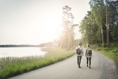 Rear view of wonderlust couple walking on road by lake against clear sky