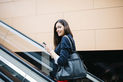 Portrait of smiling young woman using mobile phone while standing on escalator