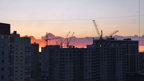 Cranes in city against sky during sunset