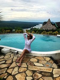 Full length of woman sitting on retaining wall by swimming pool against sky