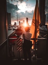 Rear view of man standing in city against sky during sunset