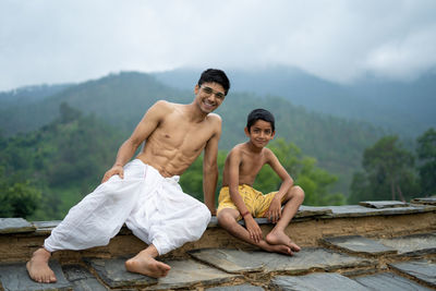 A young man sitting on the roof with his younger brother,both wearing dhoti, smiling into the camera