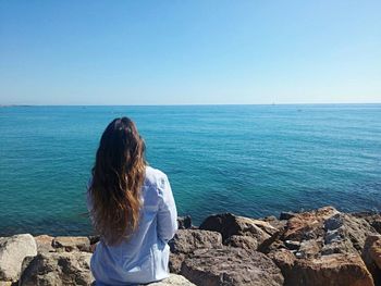 Rear view of woman sitting on rocky shore against blue sky