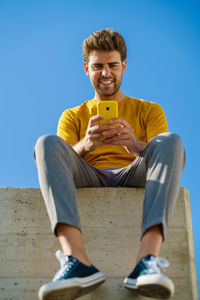 Low angel view of smiling young man using smart phone sitting against sky