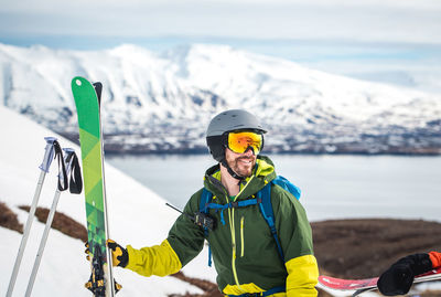 A man with backpack, radio, and skis with mountains behind him