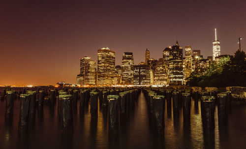 Modern illuminated buildings by lake with wooden posts against sky at night