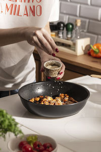 Woman adding sesame seeds to frying pan with teriyaki chicken