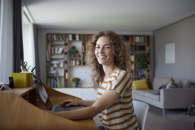 Smiling woman working on laptop while sitting by desk at home