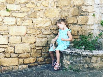 Full length of girl holding stuffed toy while sitting on retaining wall
