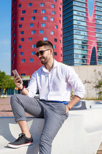 Young bearded man sits on bench outdoor against modern glass building