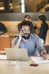 Businessman writing in diary while talking on smart phone in office