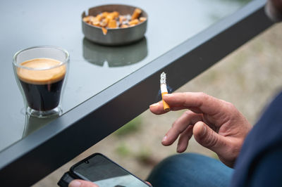 A man using smartphone while smoking cigarette and have cup of black coffee set next to him.