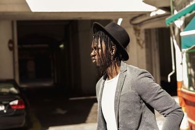 Young man with braided hair wearing hat standing against built structure