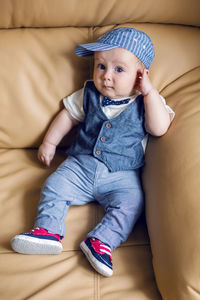 Gay boy kid blonde in a cap , pants, tie and vest sits on a light leather sofa