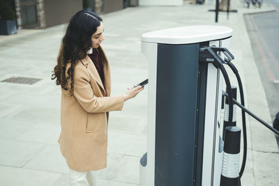 Young woman with smart phone making contactless payment at electric vehicle charging station