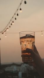 Close-up of hand holding glass of water against sky during sunset