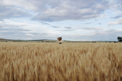 Back view of anonymous female with flying hair running on meadow with wheat spikes under cloudy sky