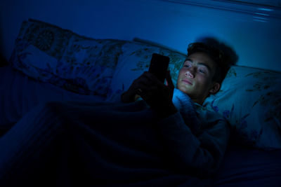 Smiling boy using smart phone while lying down on bed at home