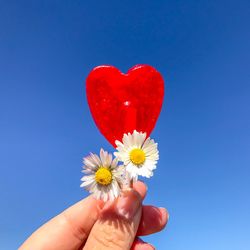 Cropped hand holding red heart shape lollipop with flowers against clear blue sky