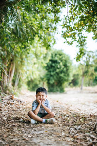 Portrait of cute boy with hands on chin sitting against trees