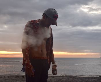 Man exhaling smoke while standing at beach against sky during sunset