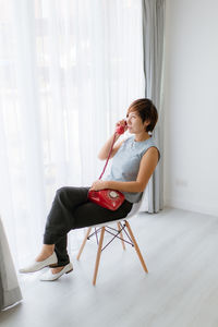 Woman using rotary phone while sitting on chair by window at home