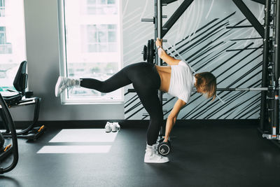 A sporty girl holding a dumbbell in her hand bends over with her in the gym, side view