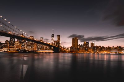 Illuminated brooklyn bridge over east river in city against sky at night