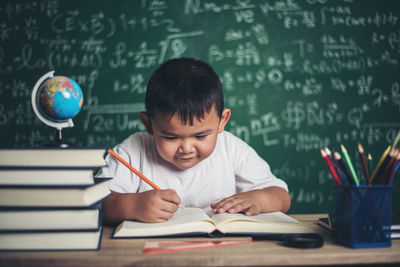 Cute boy studying at table against blackboard