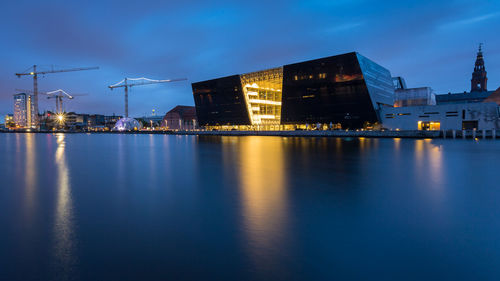 Scenic view of lake by illuminated buildings against cloudy sky at dusk